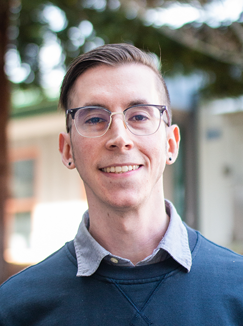 Travis Caskey smiling, standing in front of white building with tree branches in background