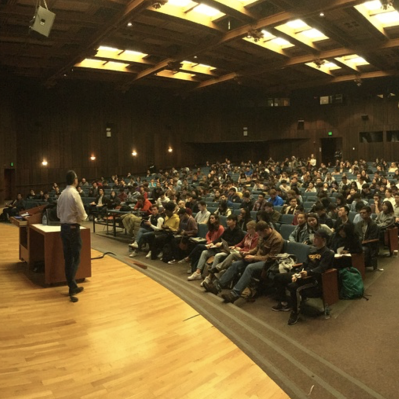 A professor lectures from a stage to a large sea of students in Data 100