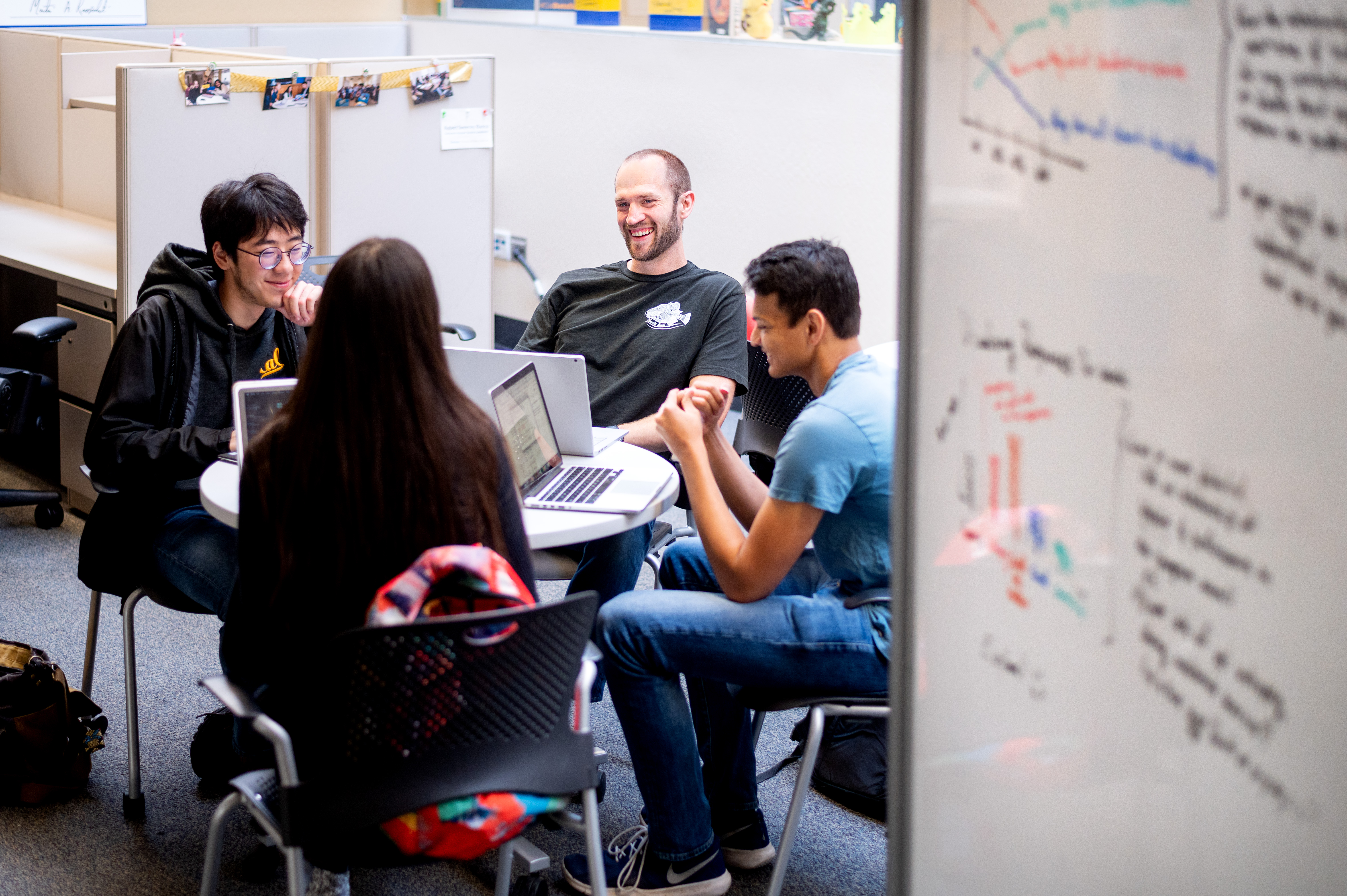 Students laugh as they collaborate around a small table, laptops open