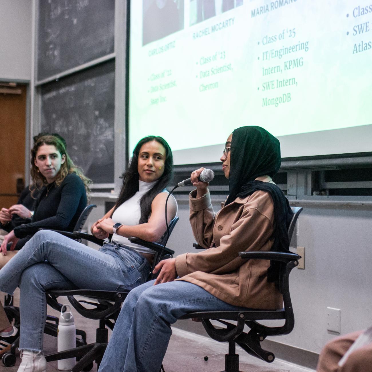 Seated panelist speaks into mic, while other panelists onstage listen.