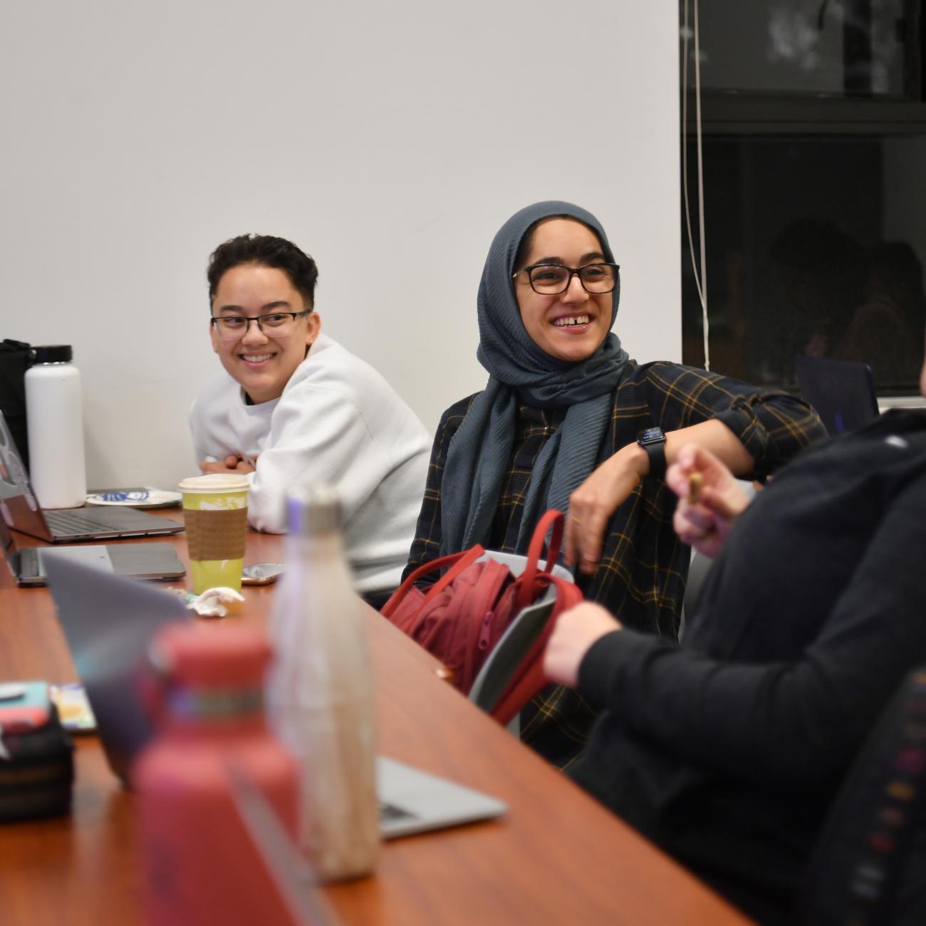 Over the shoulder of a relaxed individual turned away from the camera to talk to students seated alongside her at a table, two students listen and laugh.