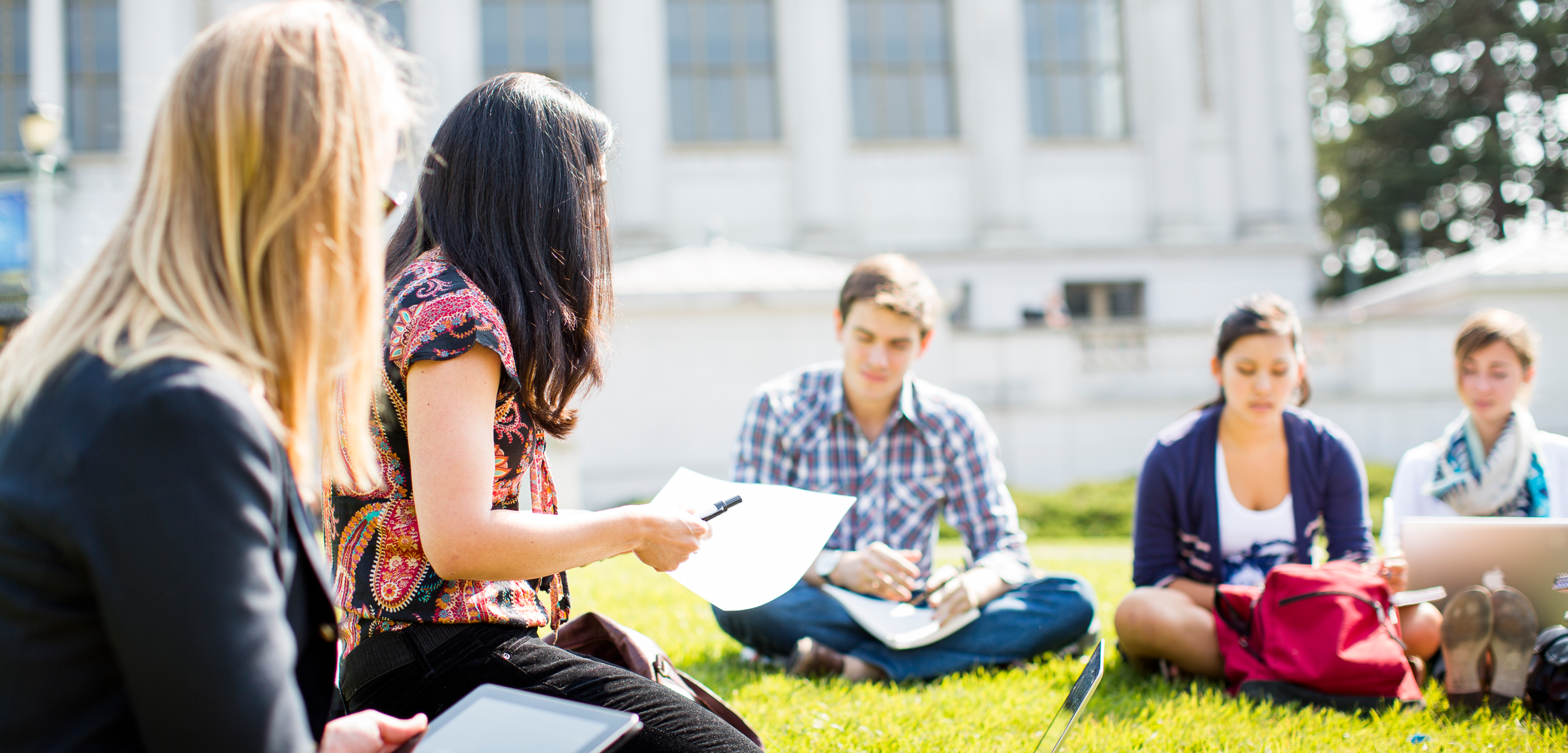 Five students sitting on grass in front of Doe Library with laptops 