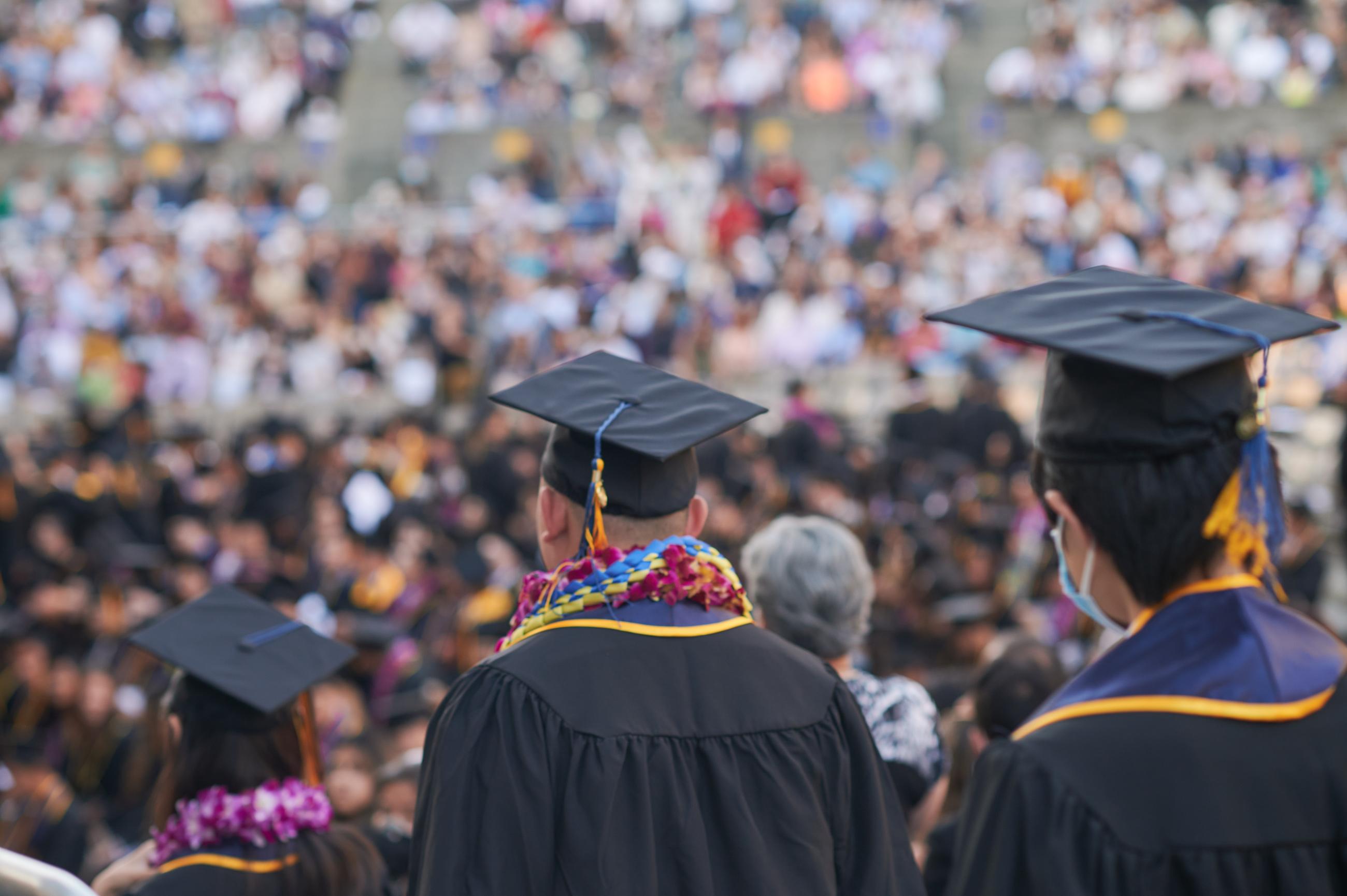 Students in graduation robes and caps walk down into stadium crowded with onlookers