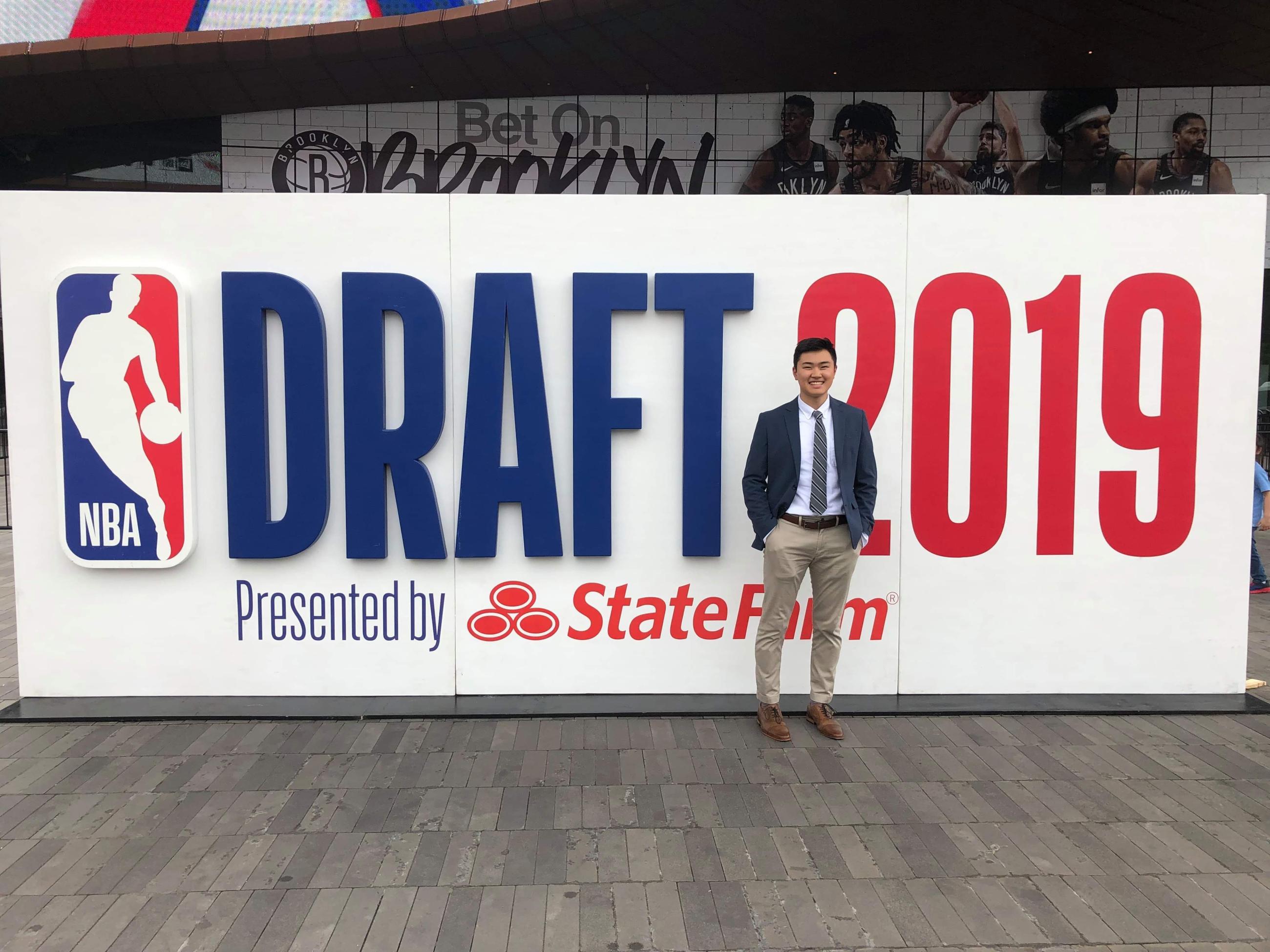 Alex Nakagawa, dressed in business attire and smiling, stands in front of the "NBA Draft 2019" sign